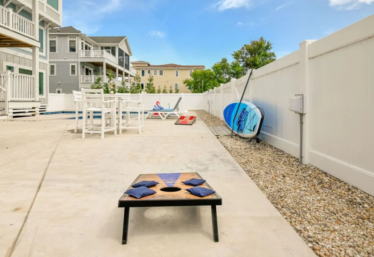 An exterior view focused on a playful outdoor area featuring a cornhole setup, surfboards, lounge chairs, and a glimpse of the pool. The scene is bright and playful, inviting guests to unwind, play games, or sunbathe by the pool.