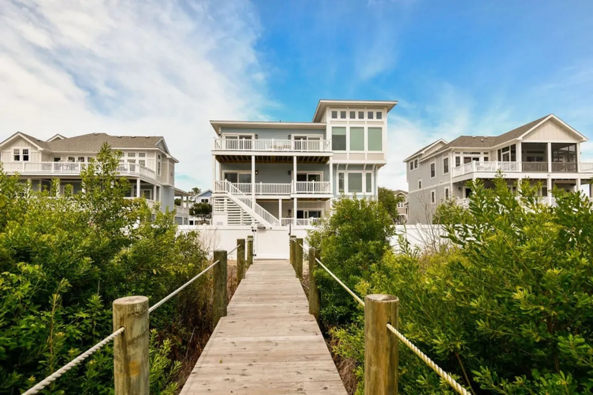 This image features the main house with a welcoming wooden walkway leading through lush greenery, setting a tranquil and inviting scene just steps from the beach. The porch is visible, hinting at relaxing mornings or evenings after a day by the ocean.