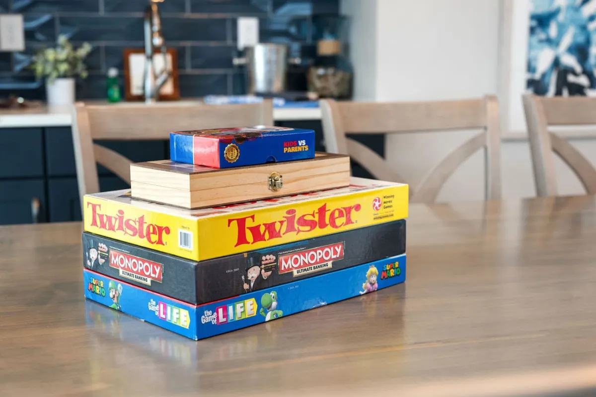 A playful, inviting scene with a stack of board games on a dining table and the kitchen in the background, suggesting a space designed for family game nights and casual gatherings.