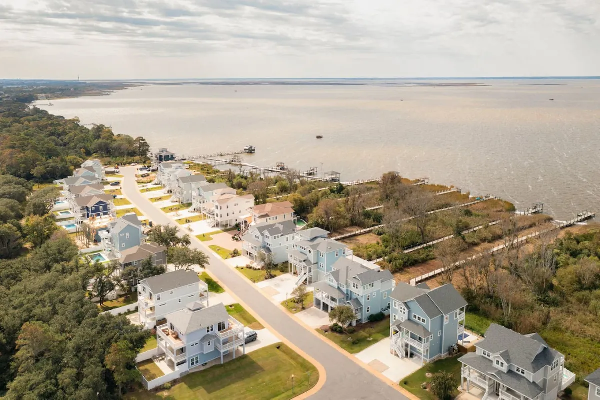 A tranquil waterfront scene featuring colorful houses that line the shore, a winding pathway, lush treeline, and expansive water views. The atmosphere is serene and welcoming, ideal for walking, relaxing by the water, or participating in boating and fishing activities.