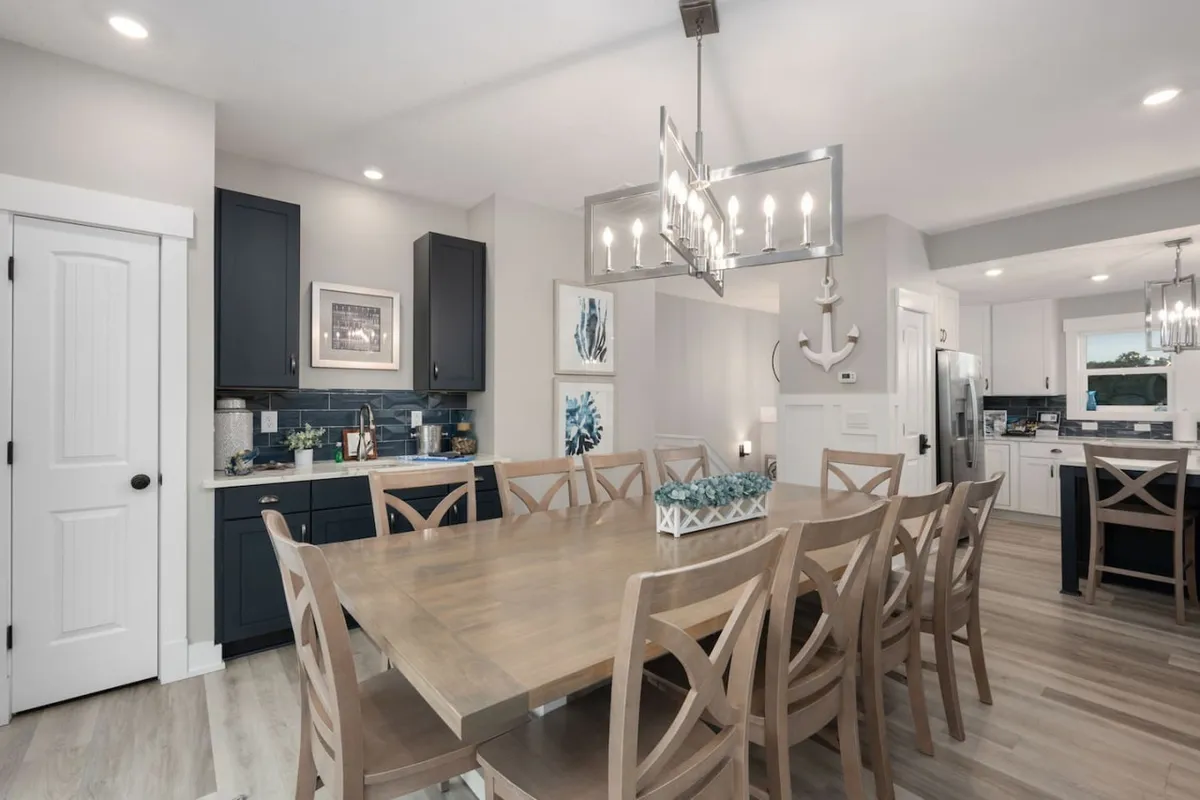 An inviting dining room with a large table set for a group, a striking chandelier overhead, navy blue cabinetry, and tasteful artwork adorning the walls. The space is perfect for hosting gatherings, dinner parties, or family meals, blending elegance with comfort.