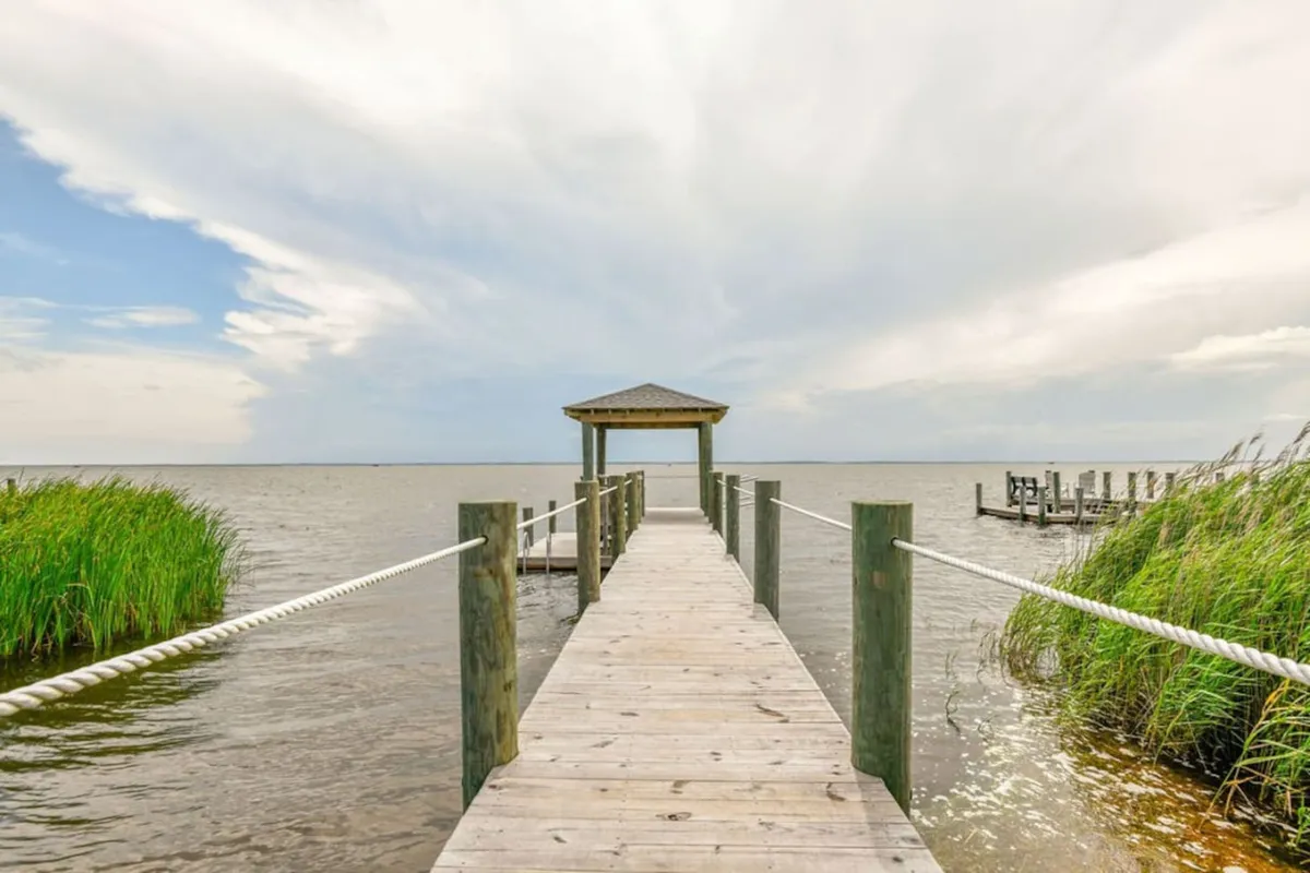 A tranquil exterior view featuring a wooden dock stretching out over calm waters, with a charming gazebo at the end and lush vegetation surrounding the scene. The open water, gentle sky, and natural greenery evoke a sense of peacefulness and direct access to the sound.