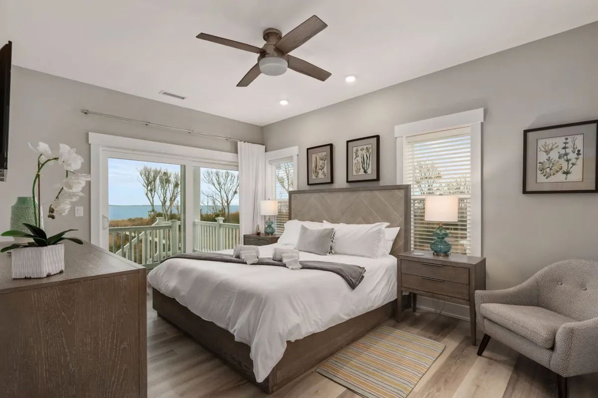 This image shows a king-size bed with plush white linens, flanked by nightstands and decorative artwork. Large windows allow plenty of natural light to fill the space, and an armchair provides an additional spot for relaxation. The tranquil setting and tasteful décor emphasize comfort and serenity.