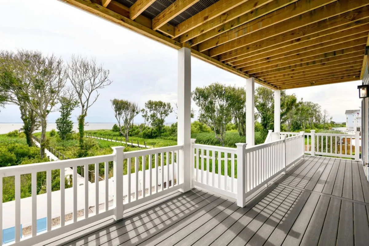 An open deck with wooden railings overlooks a sweeping expanse of greenery and a distant ocean view. Ample space for relaxing, sunset watching, or hosting outdoor gatherings is evident—an ideal spot to soak in salt air and coastal vistas.
