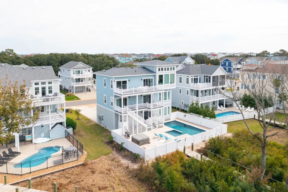 This lively exterior shot captures a swimming pool surrounded by deck areas and colorful houses in the background, all framed by well-maintained greenery. The scene exudes a sense of fun and invitation, with plenty of space for swimming, lounging, and gathering outdoors.
