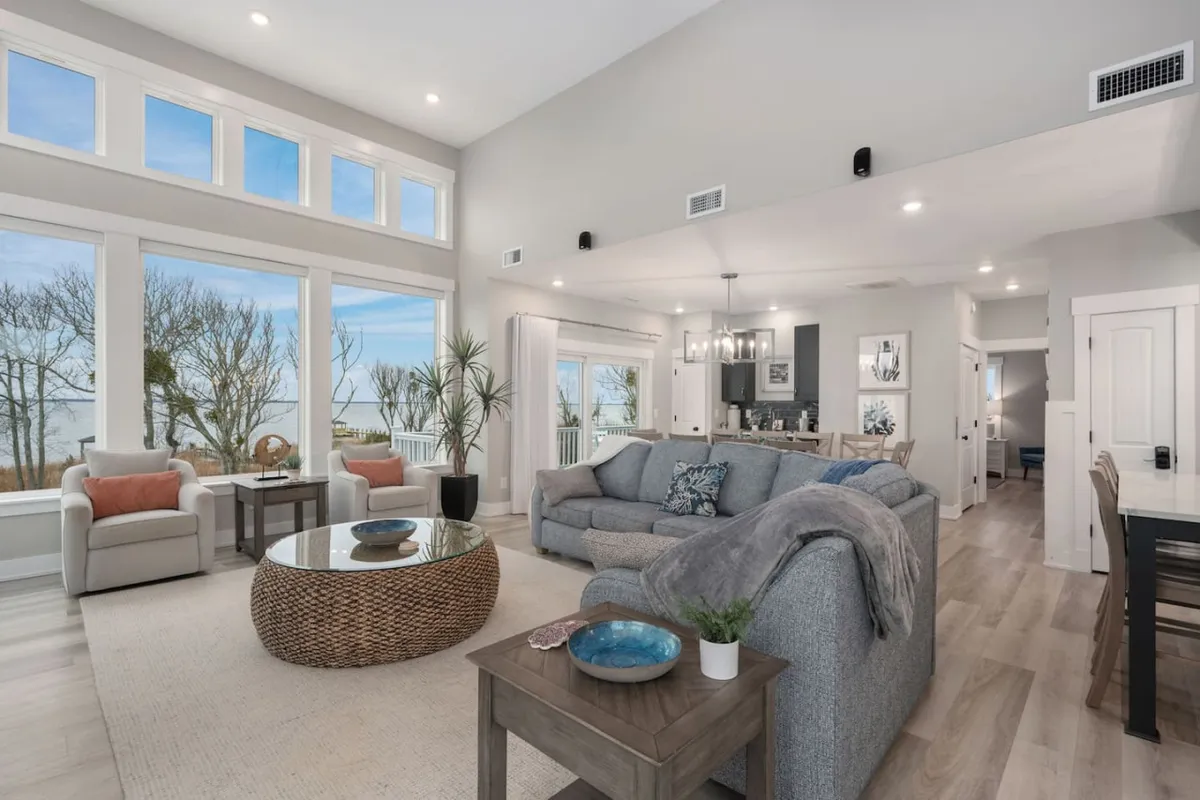 A welcoming living room with large windows bringing in natural light, a plush gray sofa, coffee table with a wicker base, and a dining area in the background. The space is designed for relaxation, conversation, and group activities.