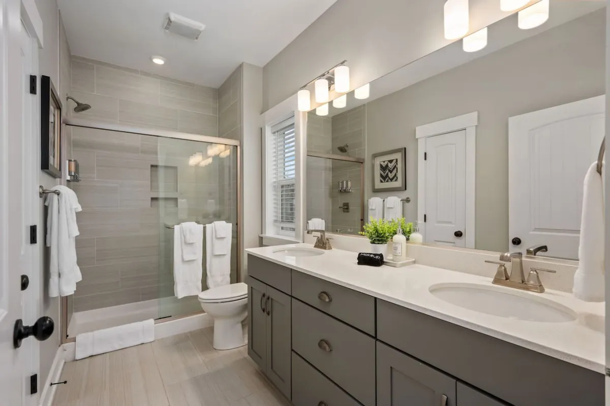 A spa-inspired bathroom featuring a double vanity sink, modern glass shower enclosure, decorative wall art, and touches of greenery on the countertop. The styling evokes a sense of calm and pampering, ideal for self-care routines after a long day.
