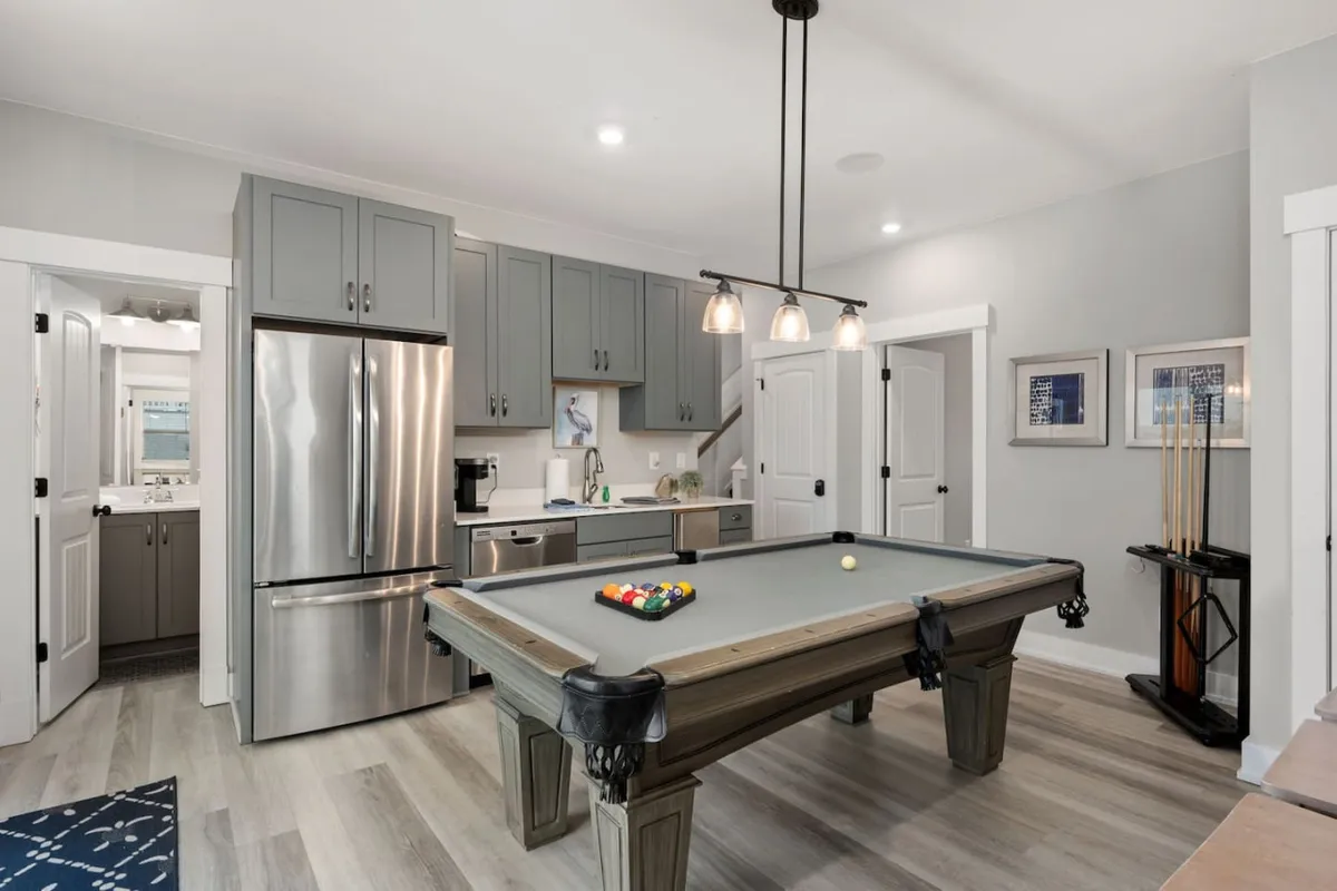 A vibrant game area featuring a pool table at the center, surrounded by kitchen appliances and tasteful lighting fixtures. Artwork adorns the walls, emphasizing the room's lively and fun atmosphere, ideal for socializing and entertainment.