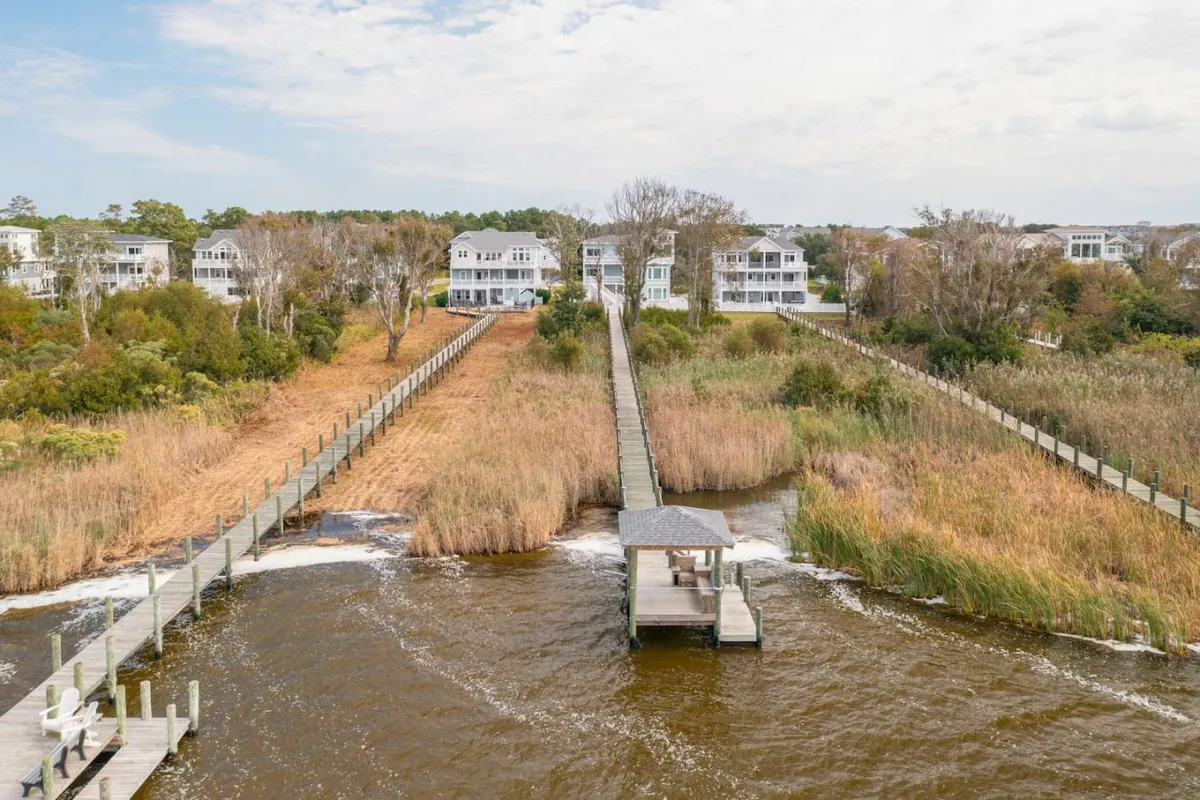A picturesque exterior view showing a boardwalk and dock extending over the water, flanked by wetland vegetation and houses in the distance. The scene highlights opportunities for boating, fishing, and relaxation amidst tranquil surroundings.