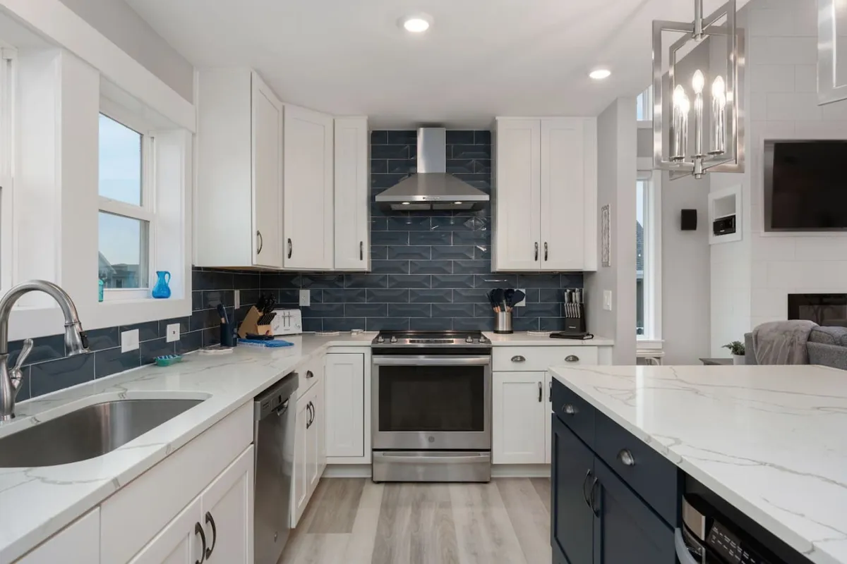 A modern kitchen featuring stainless steel appliances, a blue tile backsplash, white cabinetry, and a large island with seating, designed for both meal preparation and social gatherings. The layout exudes a contemporary and welcoming atmosphere, with ample space for cooking and entertaining.